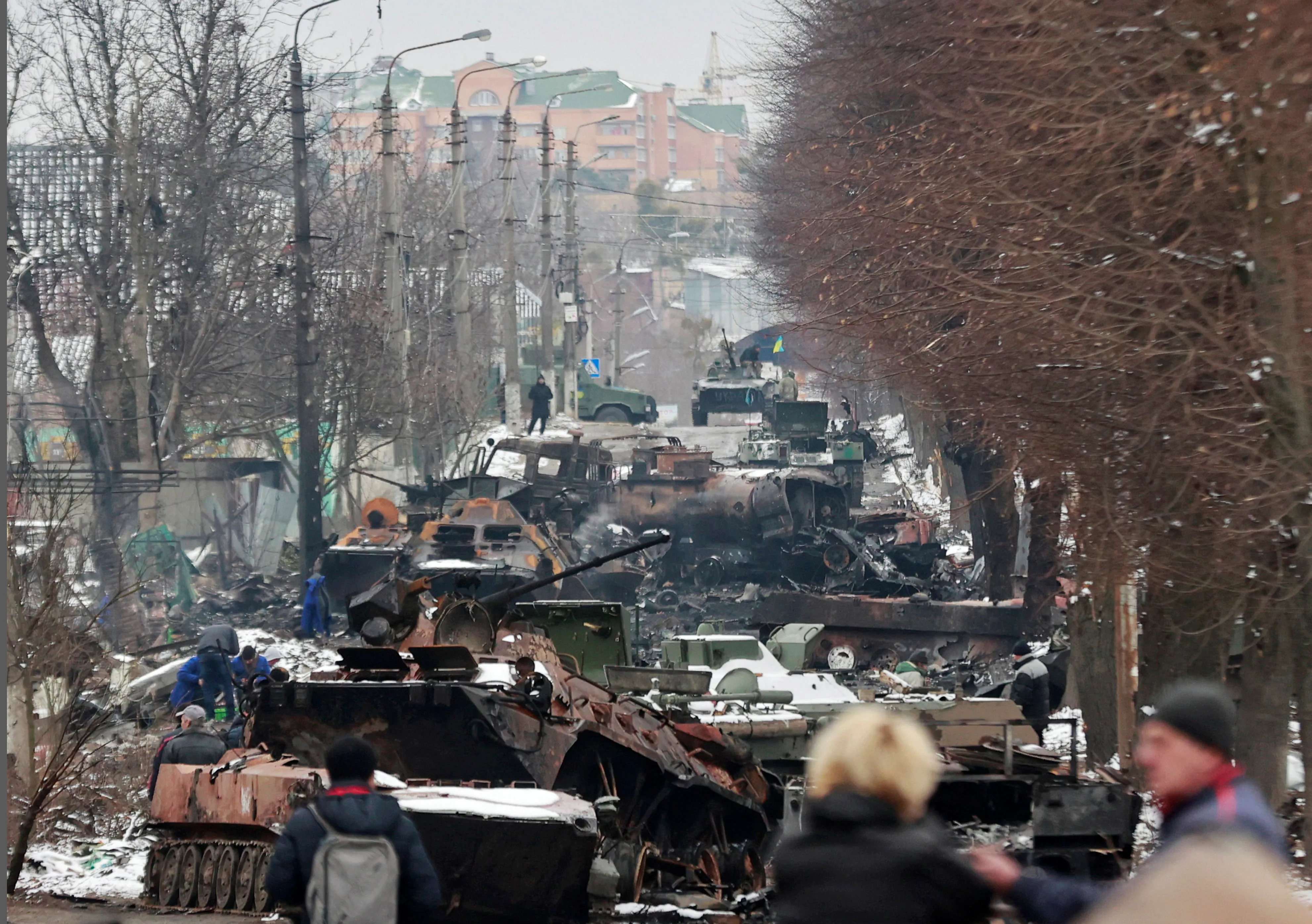 Destroyed Russian military vehicles on a street in Bucha. Civilians walk past burned-out armored personnel carriers and trucks amid snow.